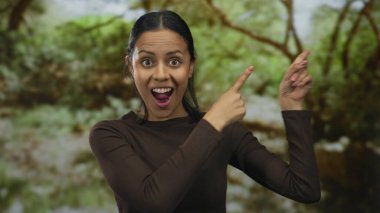 Genç İspanyol kadın yemyeşil bir açık hava parkı ortamını işaret ediyor ve gülümsüyor, doğa arasında bir heyecan ve neşe anı yakalıyor..