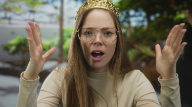 Gözlüklü kadın, orman yaprakları arasında kafasına altın bir taç takmak için iki elini kaldırır..