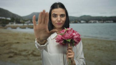 Deniz kıyısında, arka planda görünen okyanus ve dağlarla dolu bir sahilde buket tutarken el kol hareketi yapan bir kadın, dış mekan ortamında tavır ve doğa sergiliyor..