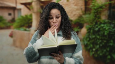 Güneşli bir sokakta kitap okuyan bir kadın yeşil bitkilerle çevrili, gözlüklü ve rahat bir kazak giymiş, derin bir düşünceye dalmış, kentsel bir eğitim anını tasvir ediyor..