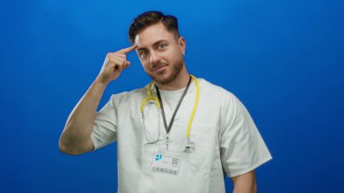 Kafasında sakal ve steteskop olan genç bir doktor canlı mavi arka plana karşı güvenli bir şekilde izole edilmiş, düşünceli ve tıbbi profesyonelliği iletiyor..