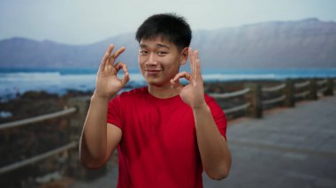 Kırmızı tişörtlü genç Çinli bir adam deniz kenarında sakin bir sahilde ve arka planda dağlarla hareket edebiliyor. Rahatlığı ve dışarıdaki sükuneti somutlaştırıyor..