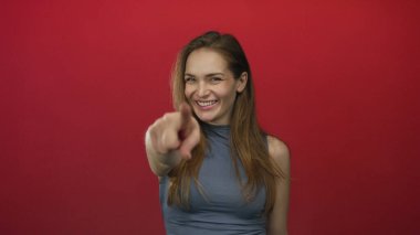 Genç beyaz kadın işaret parmağını doğrudan kameraya doğrultuyor. Parlak kırmızı stüdyo ortamında..
