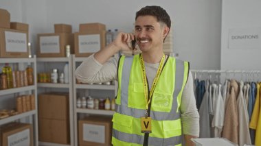 Yansıtıcı yelek giyen genç İspanyol adam kapalı bir hayır kurumunda telefonla konuşuyor. Etrafı kutu ve giysi raflarıyla çevrili. Gönüllü işleri ve sosyal yardımları resmediyor..