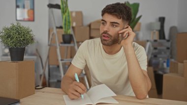 Telefondan kulağa konuşan ve bir binanın içinde karton kutularla çevrili bir masada deftere yazan bir adam, odaklanmış planlama.