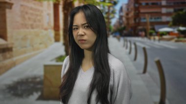 Uzun siyah saçlı, güneşli bir günde şehir sokaklarında dikilen genç Çinli kadın arkasında modern binalar olan düşünceli bir yüz ifadesi sergiliyor..