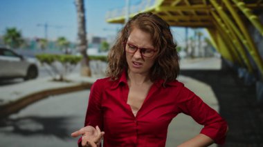Kırmızı tişörtlü kadın, modern mimari ve palmiye ağaçları tarafından çevrelenmiş güneşli şehir arka planında duygu ve hareketi yakalayan güneşli şehir caddesinde ifade veriyor..