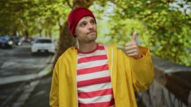 Kırmızı bereli ve sarı ceketli genç adam şehir sokağında duruyor, baş parmağıyla aşağı işaret ediyor, arka planda canlı yeşil ağaçlar ve park edilmiş arabalarla çevrili..