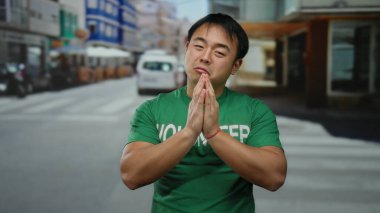 Yeşil gönüllü gömleği giyen genç adam şehir sokağında duruyor ve arkasında renkli binalar olan bir dua jesti gösteriyor..