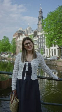 Yeşil kanalların yanında duran, gözlüklü ve beyaz kazaklı bir kadın güneşli bir günde resimsel şehir manzarasını sergiliyor..