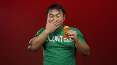 Gönüllü gömleği giyen genç adam kırmızı arka planda turuncu kurdele tutuyor..