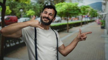 Sakallı İspanyol bir adam, şehir sokağında gülerken gündüz vakti arka planda ağaçlarla arama hareketi yapıyor..