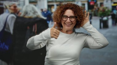 Şehir sokaklarındaki orta yaşlı esmer kadın, arka plandaki insanlarla baş parmak işareti yapıyor..
