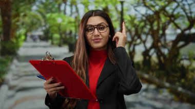 Parktaki İspanyol kadın elinde kalemle düşünceli bir şekilde kırmızı bir pano tutuyor açık hava ortamında yemyeşil bir yeşillikle çevrili iş ve doğayı harmanlıyor..