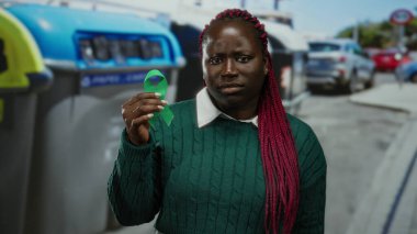 Şehir caddesinde yeşil kurdeleyi gözlemleyen bir kadın, kentsel çevre ve çeşitli elementler arasında yansıma gösteriyor..