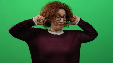Yeşil duvara yaslanmış kıvırcık saçlı, yeşil arka planda gözlüklü ve mor kazaklı bir kadın..