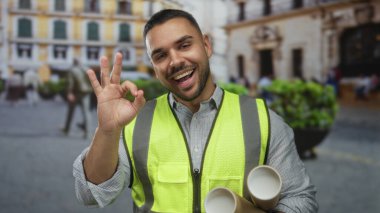 Yüksek görüşlü yelek giyen genç İspanyol mühendis, elinde yuvarlanmış mimari planlar tutarken kentin hareketli caddesinde el işareti yapıyor..