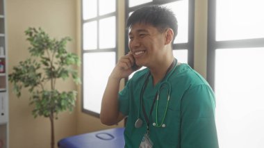 Yeşil önlüklü genç doktor gülümsüyor ve telefonda, parlak bir hastane odasında stetoskop ve kimlik kartıyla konuşuyor..