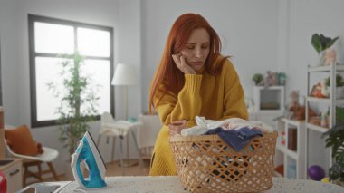 Kızıl saçlı kadın çamaşırlarını çamaşır odasında hasır sepet ve demirle temizliyor.