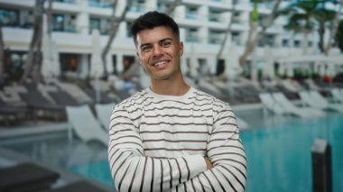 Çizgili tişörtlü genç adam, açık hava otelinde lüks bir havuz kenarında, rahat ve çekici bir tavır sergiliyor, parlak gökyüzünün altında..