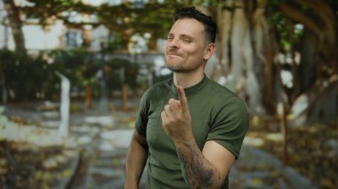 İspanyol bir adam parkta bir jest yapıyor, dövmeleri var ve yeşil bir gömlek giyiyor, doğal ortamda cazibe ve güven yayıyor..
