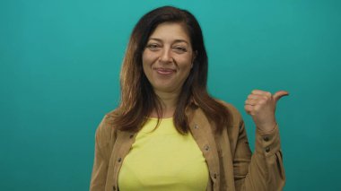 Orta yaşlı İspanyol kadın sarı gömlek ve kahverengi ceket giyiyor. Stüdyoda gülümsüyor ve el hareketi yapıyor..