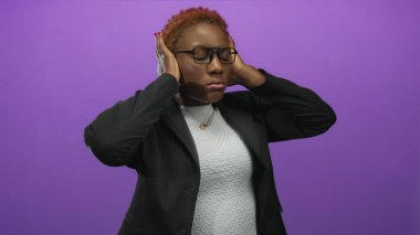 Genç Afro-Amerikalı kadın, mor stüdyoda sesi engellerken kulaklık takıyor ve parmaklarıyla kulaklarını tıkıyor..
