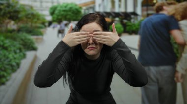 Esmer kadın bir şehir sokak dekorunda neşe içinde gözleri kaplayan, şehir yeşilliği ve açık binalar arasında şaşırmış görünüyor.