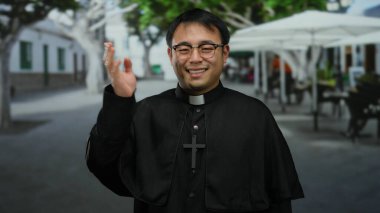 Gözlüklü ve haçlı siyah cüppeli gülümseyen rahip şehir caddesinde ağaçların ve açık hava kafelerin bulanık arka planıyla duruyor..