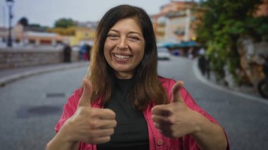 Pembe ceketli orta yaşlı kadın, şehir dışındaki dar kaldırım taşı caddesinde çifte onay gösteriyor..