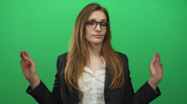 Kadın yeşil stüdyoda başını eğerek ve hafif bir gülümseme ile gözlerini kapayarak omuz silker; belirsizlik belirsizlik belirsizlik kafa karışıklığı.