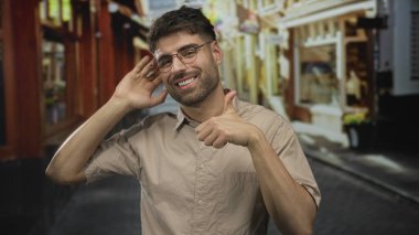 Adam el kulağına dinleme hareketi yapıyor ve güneşli kalabalık bir şehir sokağında baş parmak işareti gösteriyor; neşeli.
