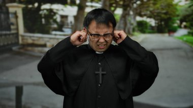 Gözlüklü ve büro kıyafeti giyen bir adam sessiz bir sokakta dikiliyor, elleri kulaklarını haç kolyesiyle kapatıyor, sıkıntı ve hüsranı iletiyor..