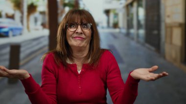 Kırmızı süveterli ve gözlüklü İspanyol bir kadın şehir sokaklarında ellerini sallayarak şehir ortamında duygularını ifade ediyor..