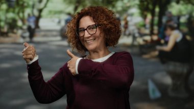 Kıvırcık esmer saçlı ve gözlüklü İspanyol bir kadın. Neşeli bir parkta güneşli bir günün tadını çıkarırken neşe içinde parmaklarını havaya doğrultuyor..