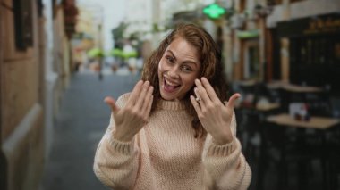 Elleri şehir terasında gezen, rahat bir kazak giyen, neşeli görünen ve şehir arka planına karşı kurulmuş açık bir kafede eğlenen bir kadın..