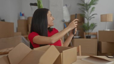 Karton kutular ve bitkilerle çevrili yeni evinde bavulunu açarken gülümseyen kadın, rahat bir oturma odasında yeni bir başlangıç ve heyecanı yansıtıyor..