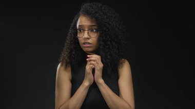 Kıvırcık saçlı bir kadın siyah bir arkaplan karşısında endişeli görünüyor, el hareketleriyle duygularını aktarıyor ve izole bir stüdyo ortamında kendini ifade ediyor..