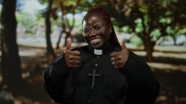 Çapraz gülümseyen ve parkın düzeninde baş parmak işareti yapan, gözlüklü ve siyah cüppeli bir kadın, doğal çevrede pozitif hava yayıyor..