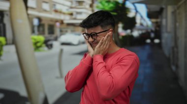 Gözlüklü, kırmızı kazaklı genç adam şehir arka planında bulanık bir sokakta duruyor. Düşünceli bir duyguyu ifade ediyor. Yüzü ellerinde dinleniyor..