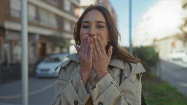Güneşli şehir caddesinde hava öpücüğü veren bir kadın bej renkli trençkotlu, neşeli bir ifade ve şehir arka planına sahip, açık hava sıcaklığını kucaklayan ve canlı bir gülümseme takınıyor..