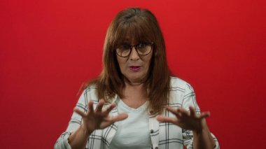 Kıdemli İspanyol kadın, canlı kırmızı bir duvara karşı sakince el kol hareketi yapıyor. Gözlüklü ve ekose gömlekli, orta boylu saçlı, kapalı alanda izole edilmiş, düşünceli bir sahne yaratıyor..