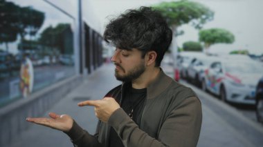 Sakallı genç İspanyol bir adam park halindeki arabaları ve camlarında yansıyan ağaçlarla bir sokakta avucunun içini işaret ederek merakı ya da talimatı yansıtıyor.