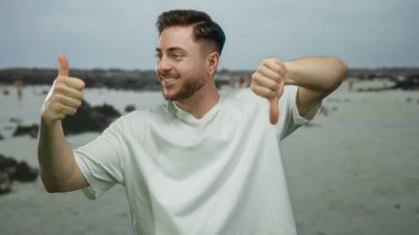 Sakallı genç adam deniz kenarındaki kumsalda baş parmağını kaldırır ve baş parmağını kaldırır. Kayalar ve bulutlu gökyüzü açık havada karışık duygular gösterir.