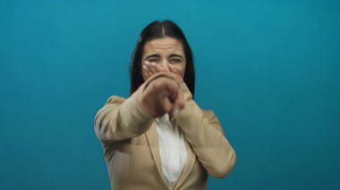 Bej takım elbiseli genç İspanyol kadın, gülüyor ve mavi bir duvar arkasına işaret ediyor, neşeli duyguları güvenli bir şekilde ifade ediyor..