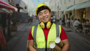 Güvenlik teçhizatı takan gülümseyen adam arka planda kafeleri olan bir şehir sokağının planlarını tutuyor..