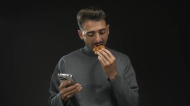 Siyah stüdyo ortamında akıllı telefon tutarken waffle yiyen adam; çoklu görev kolaylığı rahatlaması.