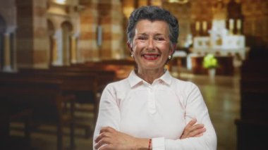 Kilise ortamında gülümseyen, kollarını kavuşturmuş, beyaz gömlek giyen, süslü mimari ve ahşap sıraları olan, dingin ve neşeli bir atmosfer yakalayan yaşlı kadın..