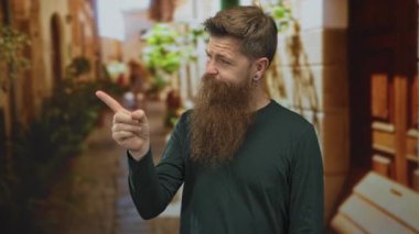 Uzun sakallı beyaz bir genç adam işaret parmağını dar, eski bir kasaba sokağında gökyüzüne doğru gösteriyor..