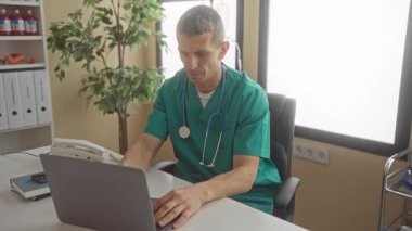 Doktor üniformalı ve steteskoplu bir adam modern sağlık ortamını gösteren klinik ofis ortamında dizüstü bilgisayarla çalışıyor..